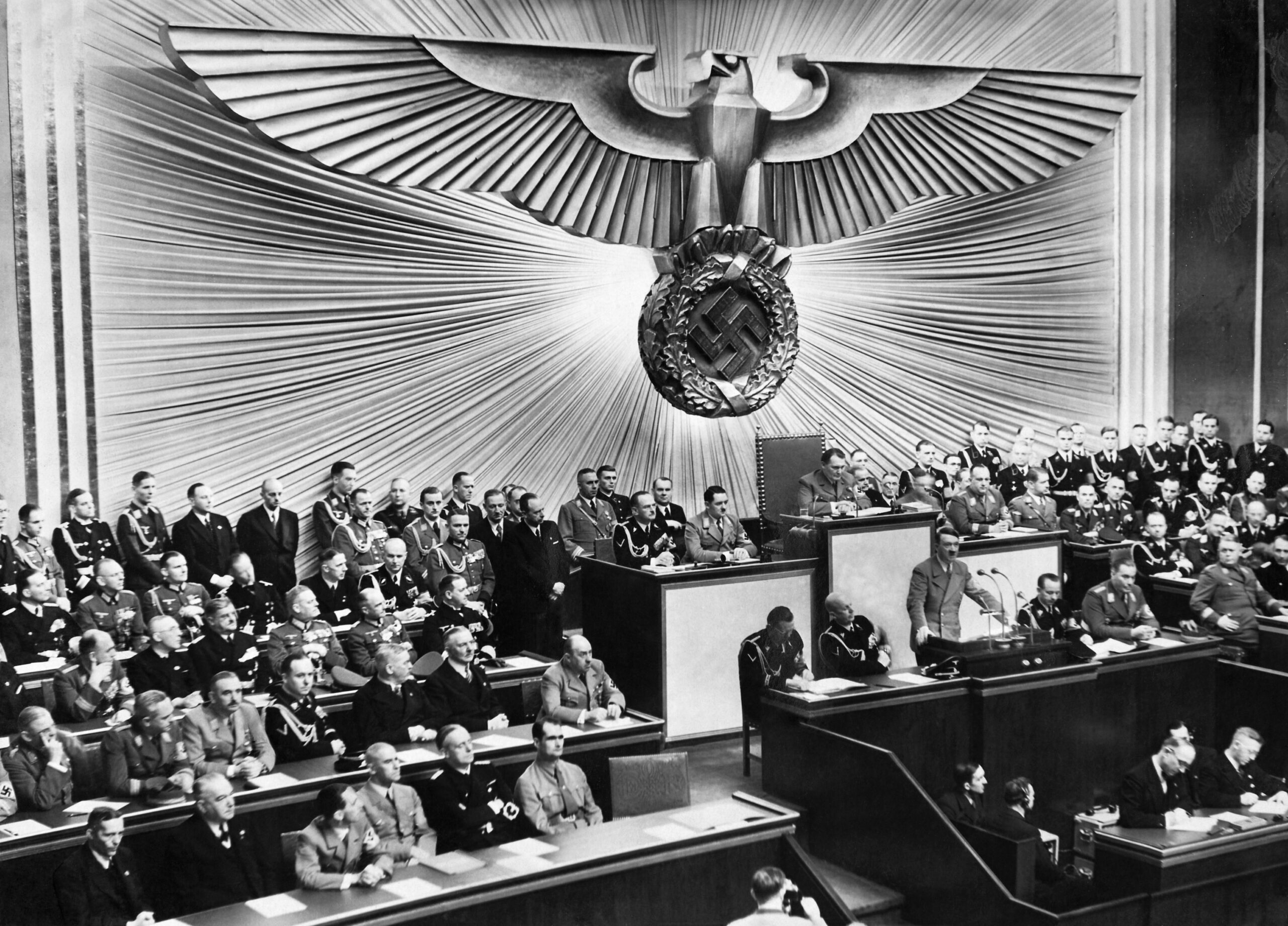 Adolf Hitler de pie ante un podio, hablando a una gran asamblea de hombres uniformados y trajeados en una cámara decorada con una gran águila con esvástica.