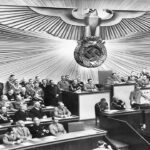 Adolf Hitler de pie ante un podio, hablando a una gran asamblea de hombres uniformados y trajeados en una cámara decorada con una gran águila con esvástica.