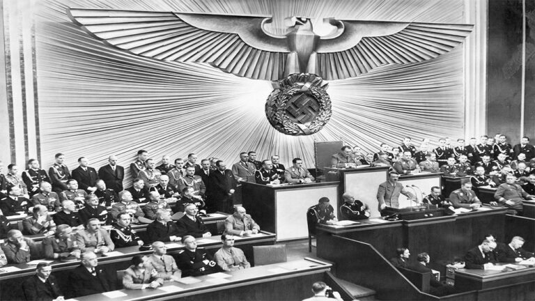 Adolf Hitler de pie ante un podio, hablando a una gran asamblea de hombres uniformados y trajeados en una cámara decorada con una gran águila con esvástica.