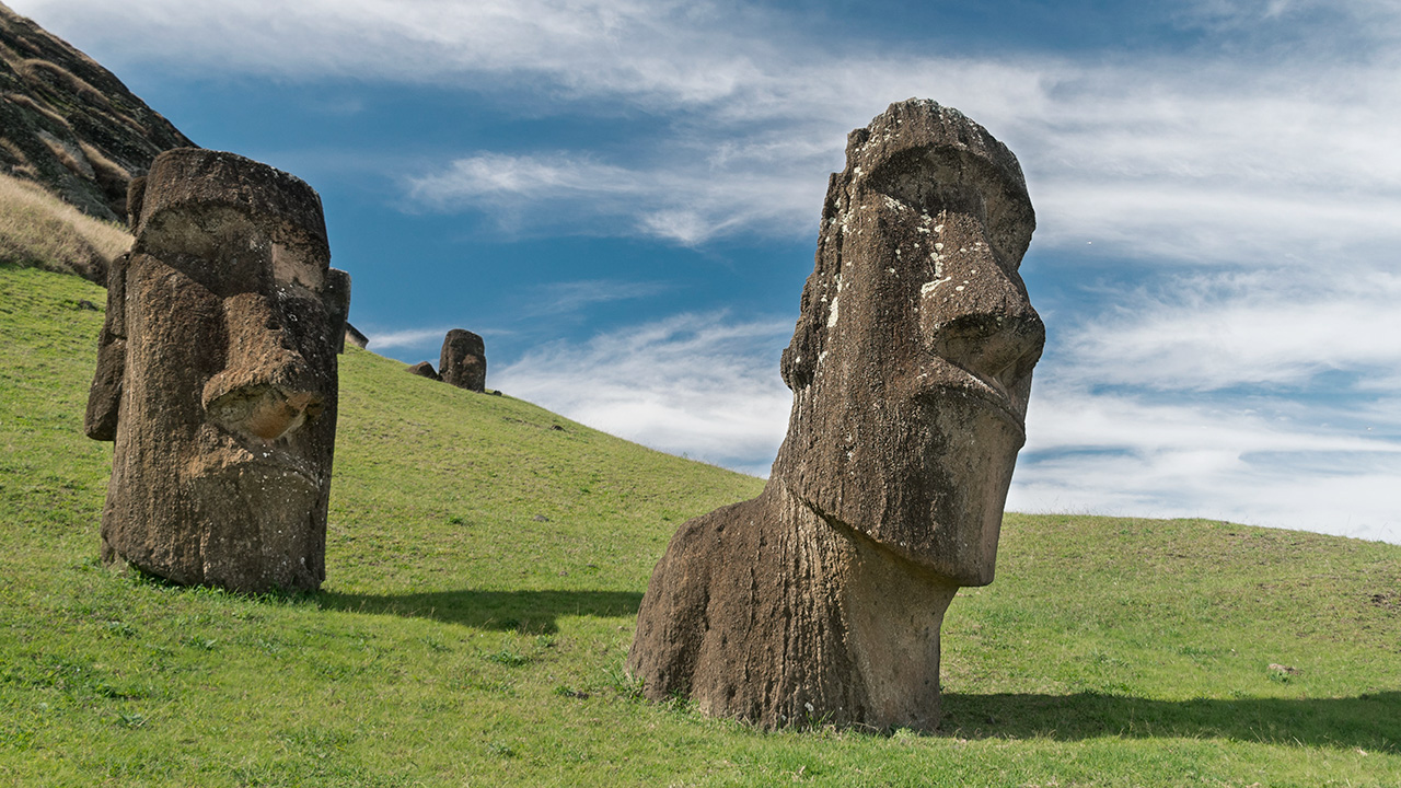 Plano medio de dos imponentes estatuas de piedra Moái parcialmente enterradas en la ladera verde de Rano Raraku bajo un cielo azul con nubes ligeras.