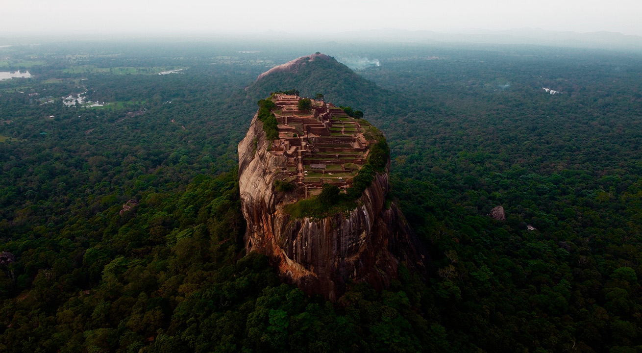 Vista aérea de la imponente roca monolítica de Sigiriya con las ruinas de la fortaleza en su cima, rodeada por una densa selva verde.