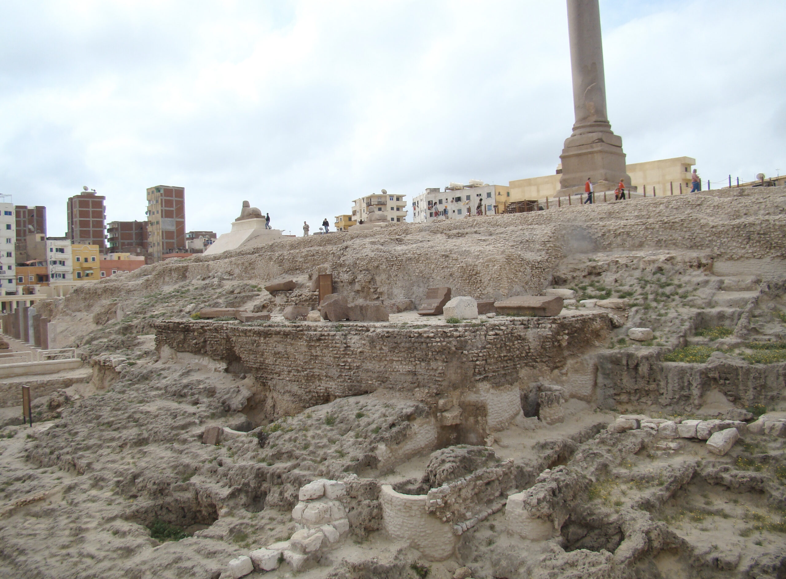 Vista panorámica de una excavación arqueológica con ruinas antiguas, la gran columna de Pompeyo, estelas de piedra labrada en primer plano y la ciudad moderna al fondo.